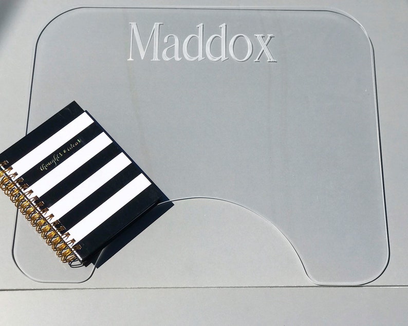 Op de afbeelding: Een transparante acryl bureaumat met de naam "Maddox" erop gegraveerd. Een zwart-wit gestreept notitieboek met een gouden spiraalbinding ligt op de mat.
