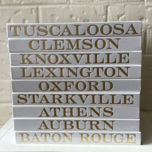 May include: A stack of white books with gold lettering. The book spines display the names of cities: Tuscaloosa, Clemson, Knoxville, Lexington, Oxford, Starkville, Athens, Auburn, and Baton Rouge. The books are stacked on a white surface.