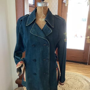 May include: A double-breasted denim jacket in a medium blue hue, displayed on a vintage dress form. The jacket features a notched collar, button closures, and decorative shoulder epaulets. The sleeves are rolled up, revealing a patterned lining.