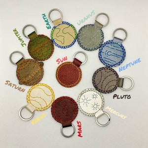May include: A set of 10 embroidered keychains featuring the planets of our solar system. Each keychain is a round, colorful patch with a different planet design and the planet's name written in cursive.