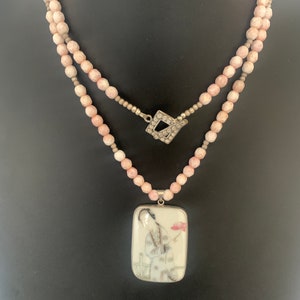 May include: A double-strand necklace with pink beads and a rectangular pendant. The pendant features a painted image of a person in traditional attire holding a flower. The necklace has a silver clasp and pendant setting.