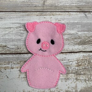 May include: Pink felt finger puppet of a pig with embroidered details. The pig has a pink snout, black eyes, and a white smile.