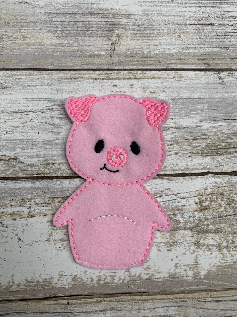 May include: Pink felt finger puppet of a pig with embroidered details. The pig has a pink snout, black eyes, and a white smile.