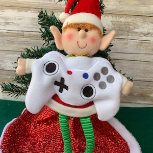 May include: A Christmas tree elf wearing a white and red Santa hat and a white shirt with a video game controller design. The elf has green and white striped legs and is sitting on a red and white tree skirt.