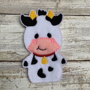 May include: A white felt finger puppet shaped like a cow with black spots, black ears with yellow tips, a pink nose, and a yellow bell around its neck.