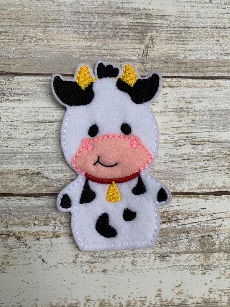 May include: A white felt finger puppet shaped like a cow with black spots, black ears with yellow tips, a pink nose, and a yellow bell around its neck.