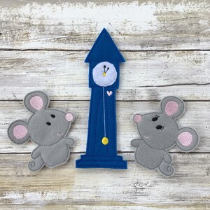 May include: Two gray felt mice with pink ears and black eyes stand on either side of a blue felt clock tower with a white clock face and a yellow pendulum.