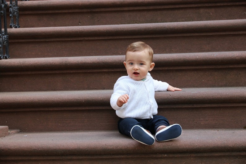 Op de afbeelding: Een baby in een witte gebreide trui, marineblauwe broek en blauwe schoenen zit op bruine traptreden. De baby kijkt met open mond naar de camera. De treden zijn gemaakt van een roodbruin materiaal.