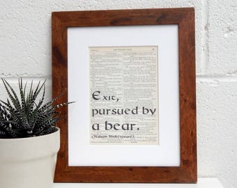 May include: A framed print of a page from a book with the text "Exit, pursued by a bear."  The text is in a black font and is surrounded by a white border. The frame is brown wood.