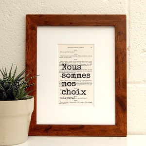 May include: A framed print of a book page with the text "Nous sommes nos choix (Sartre)". The text is in black and white and is surrounded by a white border. The frame is brown wood.