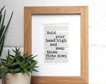 May include: A framed print of a book page with a quote from "Harper Lee" that reads "Hold your head high and keep those fists down". The print is in a light wood frame and is sitting on a table with a green plant.
