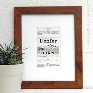 May include: A framed print of a page from a book with the text "L'enfer, c'est les autres" (Hell is other people) by Jean-Paul Sartre. The text is in black and white and the frame is brown wood.