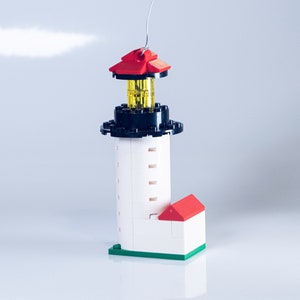 May include: A white Lego lighthouse ornament with a red roof, black base, and a yellow light. The lighthouse has a green base and a silver hook for hanging.