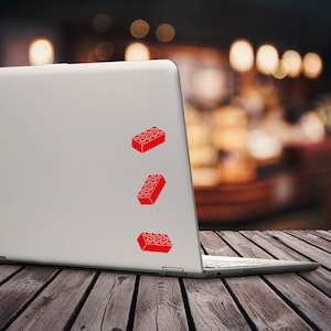 May include: A silver laptop with three red brick stickers applied to the lid. The stickers are arranged vertically, with each brick design featuring the classic studded top. The laptop sits on a wooden surface.