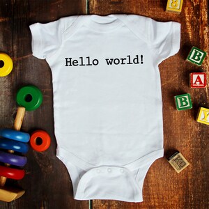 May include: White baby onesie with the text "Hello world!" printed on the front. The onesie is surrounded by colorful wooden blocks and toys.