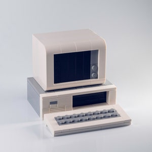 May include: A vintage computer model made of off-white and gray building blocks. The set includes a monitor, a CPU, and a keyboard. The monitor has a dark screen, and the keyboard has gray keys. The computer is a retro design.