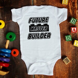 Könnte beinhalten: Weißer Baby-Strampler mit schwarzem Text "Future Master Builder" und einer schwarzen Lego-Stein-Grafik. Der Strampler ist von bunten Holzbausteinen umgeben.