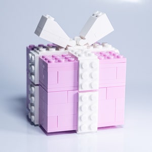 May include: A pink and white Lego gift box with a white bow on top. The box is made of plastic bricks and has a lid that opens.