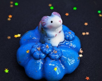 May include: A small, white, mouse-like figurine with purple-tinted ears and accents, perched atop a glossy, vibrant blue, pumpkin-shaped base. The blue base features small, sculpted flower details and is speckled with tiny, reflective gold-colored flakes.