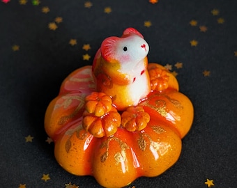 May include: A small ceramic figurine of a white and orange mouse sitting atop a glossy, bright orange pumpkin. The pumpkin is decorated with gold leaf vine patterns and features two smaller orange pumpkins resting at the base of the mouse.