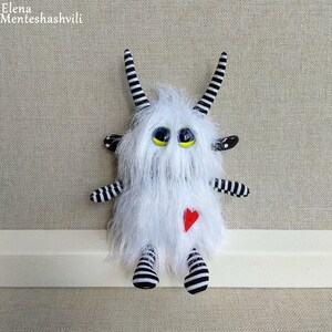 May include: White furry monster toy with black and white striped arms and legs, black and white striped horns, and large yellow eyes with black pupils. The monster is wearing a red heart on its chest.