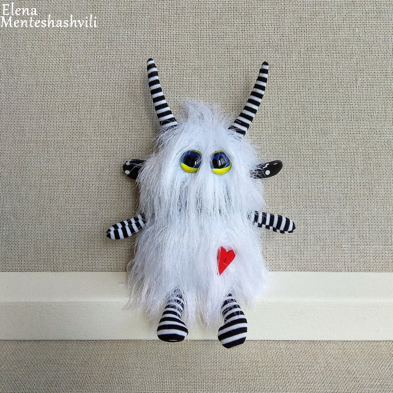 May include: White furry monster toy with black and white striped arms and legs, black and white striped horns, and large yellow eyes with black pupils. The monster is wearing a red heart on its chest.