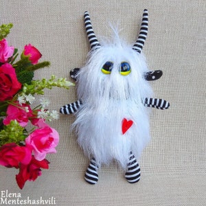 May include: A white furry toy with black and white striped arms and legs. The toy has large yellow eyes with black pupils and a red heart on its chest.