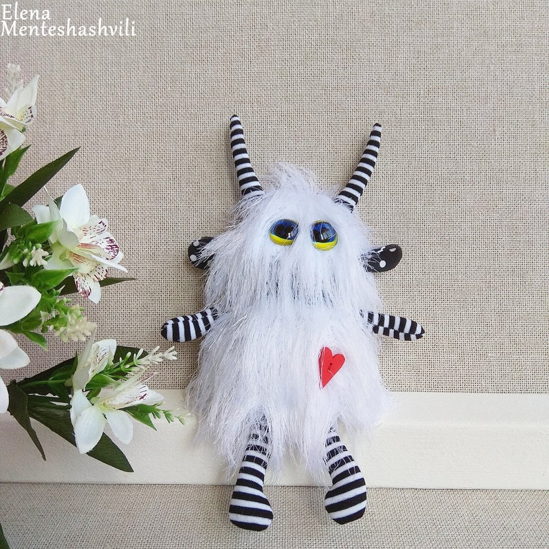 May include: White furry monster toy with black and white striped legs and arms. The monster has large yellow eyes and a red heart on its chest.