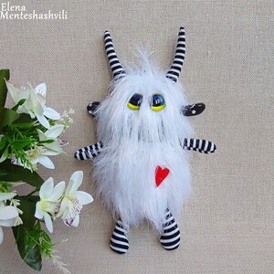May include: White furry monster toy with black and white striped arms and legs. The toy has large yellow eyes and a red heart on its chest.