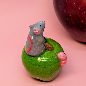 May include: A small, gray, sculpted mouse with pink inner ears and yellow star accents sits atop a glossy, bright green apple figurine. A small pink worm is attached to the side of the green apple, which features gold flecks throughout its surface.