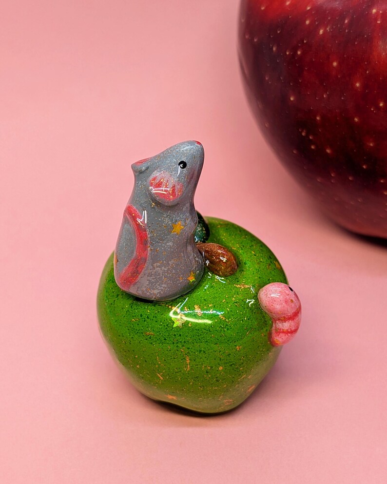 May include: A small, gray, sculpted mouse with pink inner ears and yellow star accents sits atop a glossy, bright green apple figurine. A small pink worm is attached to the side of the green apple, which features gold flecks throughout its surface.