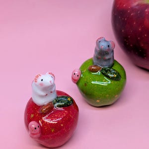 May include: Two small ceramic figurines of mice perched on glossy, glittery apples. One white mouse sits on a red apple with gold star accents, while a gray mouse sits on a green apple. Each apple features a small pink worm-like detail.