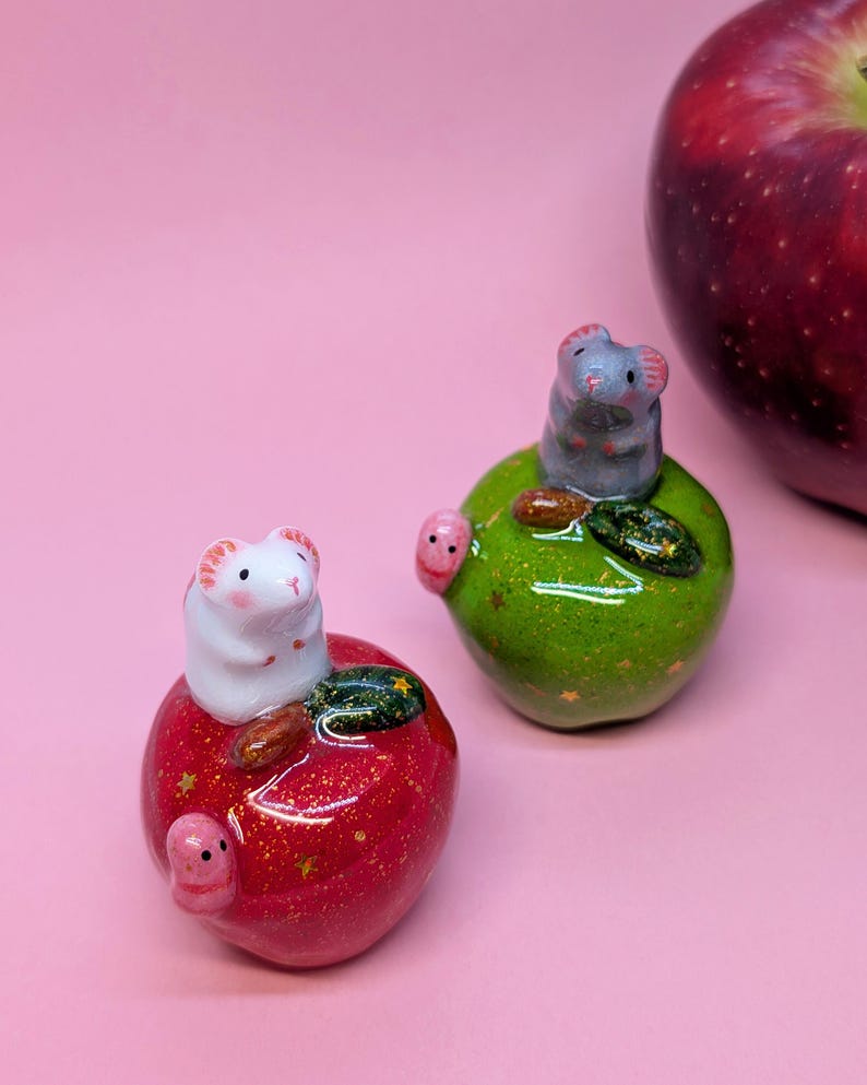 May include: Two small ceramic figurines of mice perched on glossy, glittery apples. One white mouse sits on a red apple with gold star accents, while a gray mouse sits on a green apple. Each apple features a small pink worm-like detail.