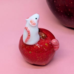 May include: A small white mouse figurine with pink-tinted ears and tail, perched atop a glossy red apple. The apple features a shiny finish with embedded gold glitter and small gold star accents, with a brown stem visible near the mouse.