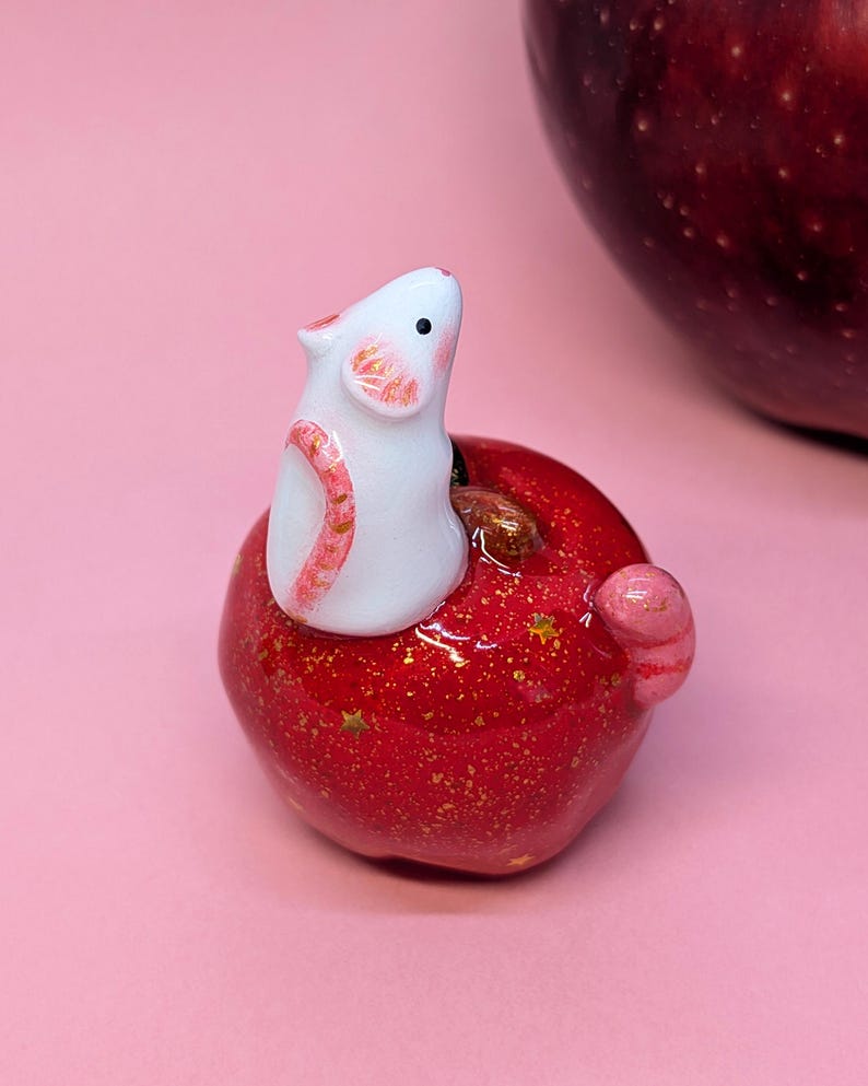 May include: A small white mouse figurine with pink-tinted ears and tail, perched atop a glossy red apple. The apple features a shiny finish with embedded gold glitter and small gold star accents, with a brown stem visible near the mouse.