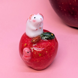 May include: A small white mouse figurine with pink ears and cheeks sits atop a glossy red apple. The apple features gold glitter, small gold star accents, a dark green leaf, and a tiny pink worm crawling on its side surface.