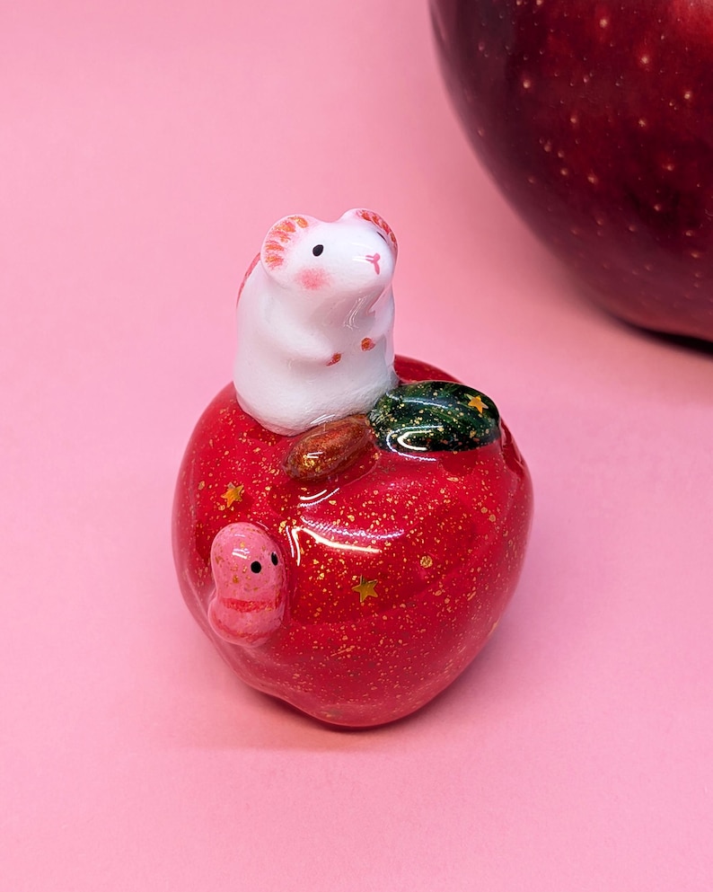 May include: A small white mouse figurine with pink ears and cheeks sits atop a glossy red apple. The apple features gold glitter, small gold star accents, a dark green leaf, and a tiny pink worm crawling on its side surface.