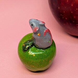 May include: A small, glossy gray mouse figurine with pink inner ears and tiny star accents, perched atop a shiny, glittery green apple sculpture that features a dark green leaf and small gold star details on its rounded surface.