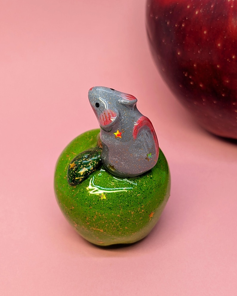 May include: A small, glossy gray mouse figurine with pink inner ears and tiny star accents, perched atop a shiny, glittery green apple sculpture that features a dark green leaf and small gold star details on its rounded surface.