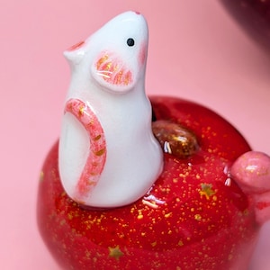 May include: A small white ceramic mouse figurine with pink-painted ears and tail, perched atop a glossy red apple-shaped object. The red surface features embedded gold glitter and small star-shaped accents, with a brown stem visible near the mouse.