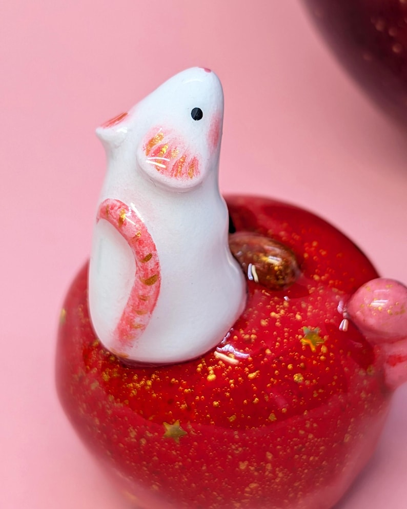 May include: A small white ceramic mouse figurine with pink-painted ears and tail, perched atop a glossy red apple-shaped object. The red surface features embedded gold glitter and small star-shaped accents, with a brown stem visible near the mouse.