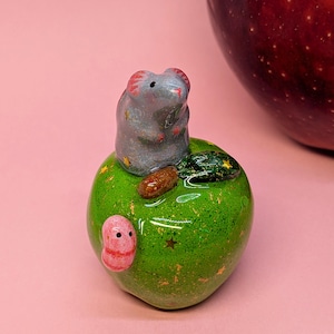 May include: A small ceramic figurine featuring a gray mouse sitting atop a glossy green apple. The apple is decorated with tiny gold star accents, a brown stem, a dark green leaf, and a small pink worm crawling on its side.