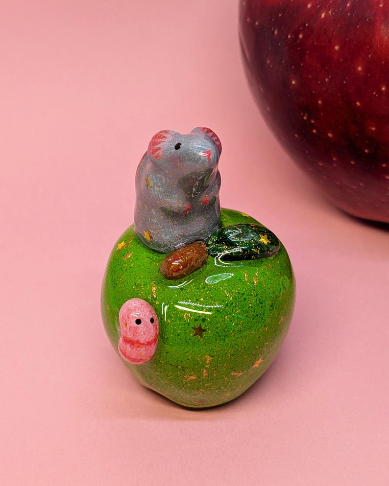 May include: A small ceramic figurine featuring a gray mouse sitting atop a glossy green apple. The apple is decorated with tiny gold star accents, a brown stem, a dark green leaf, and a small pink worm crawling on its side.