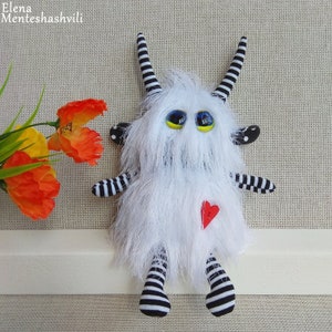 May include: White furry monster toy with black and white striped legs and arms. The monster has large blue eyes with yellow pupils and a red heart on its chest.