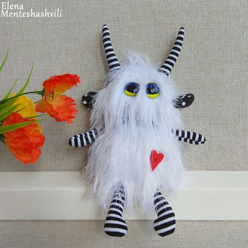 May include: White furry monster toy with black and white striped legs and arms. The monster has large blue eyes with yellow pupils and a red heart on its chest.