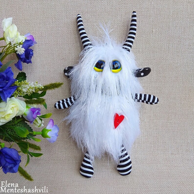 May include: White furry monster toy with black and white striped arms and legs. The monster has large blue eyes with yellow pupils and a red heart on its chest.