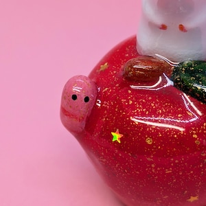 May include: A glossy red apple-shaped figurine featuring a small white creature on top, a tiny pink worm with black eyes on the side, and embedded gold glitter flakes and small holographic star-shaped accents throughout the surface.