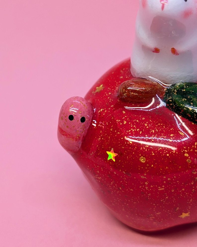 May include: A glossy red apple-shaped figurine featuring a small white creature on top, a tiny pink worm with black eyes on the side, and embedded gold glitter flakes and small holographic star-shaped accents throughout the surface.