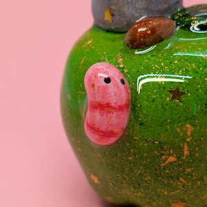May include: A glossy green ceramic object with a speckled texture and gold star accents. A smooth, pink, bean-shaped figure with two black dots for eyes is attached to the side. A brown, textured, oval-shaped stone is embedded on the top surface.