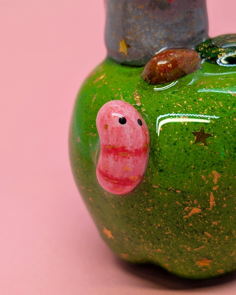 May include: A glossy green ceramic object with a speckled texture and gold star accents. A smooth, pink, bean-shaped figure with two black dots for eyes is attached to the side. A brown, textured, oval-shaped stone is embedded on the top surface.